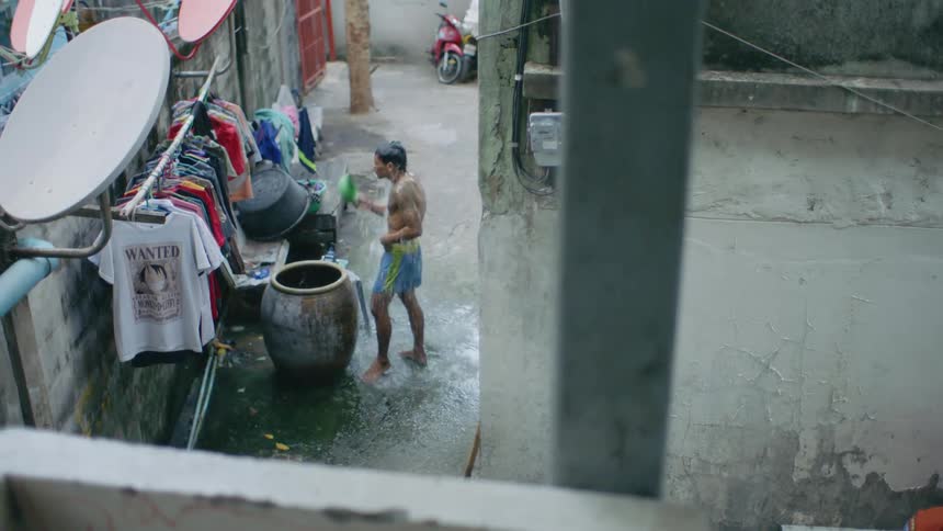 A man wearing only a pair of shorts repeatedly scoops water from a barrel with a bowl and pours it over his head to wash himself in a narrow alleyway between buildings during the day
