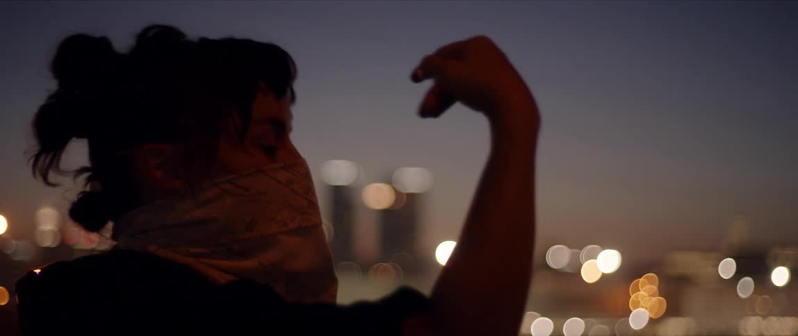 Woman with a face covering outside at night performing a dance move with her hand on the roof of a building in a city