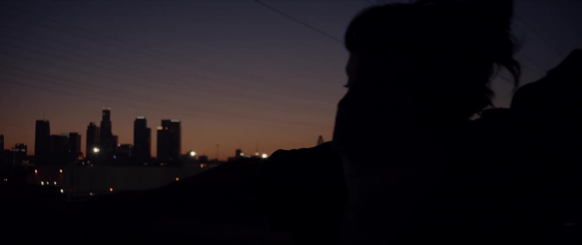 The silhouette of a young woman with her face covered by a bandana dancing on a bridge in a metropolis at night