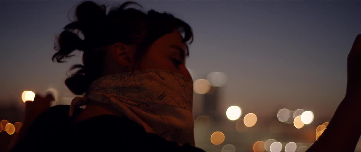 A young woman with a bandana around her lower face dances outdoors with the skyline of a metropolis at her back at night