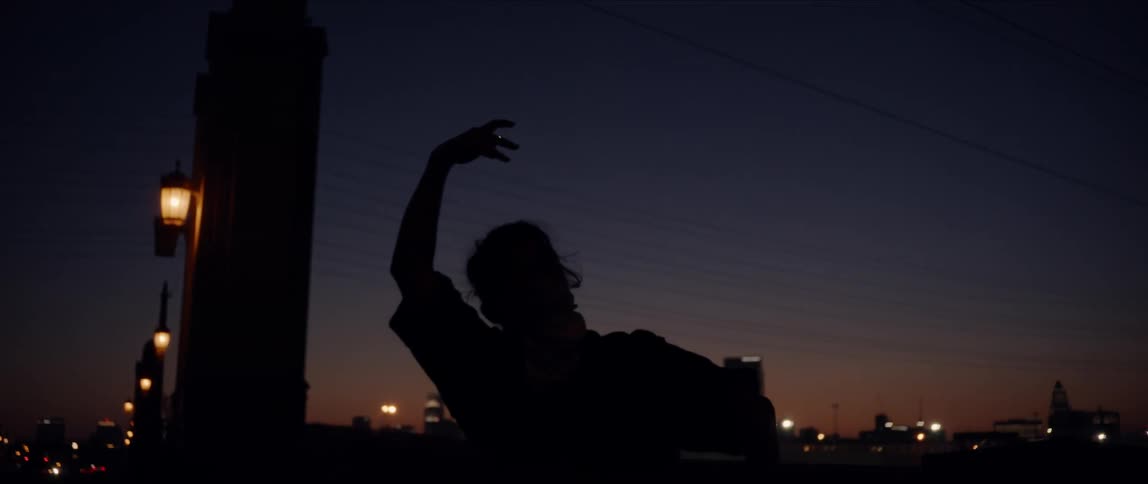 A woman performs dance moves outdoors at night with a skyline in the background