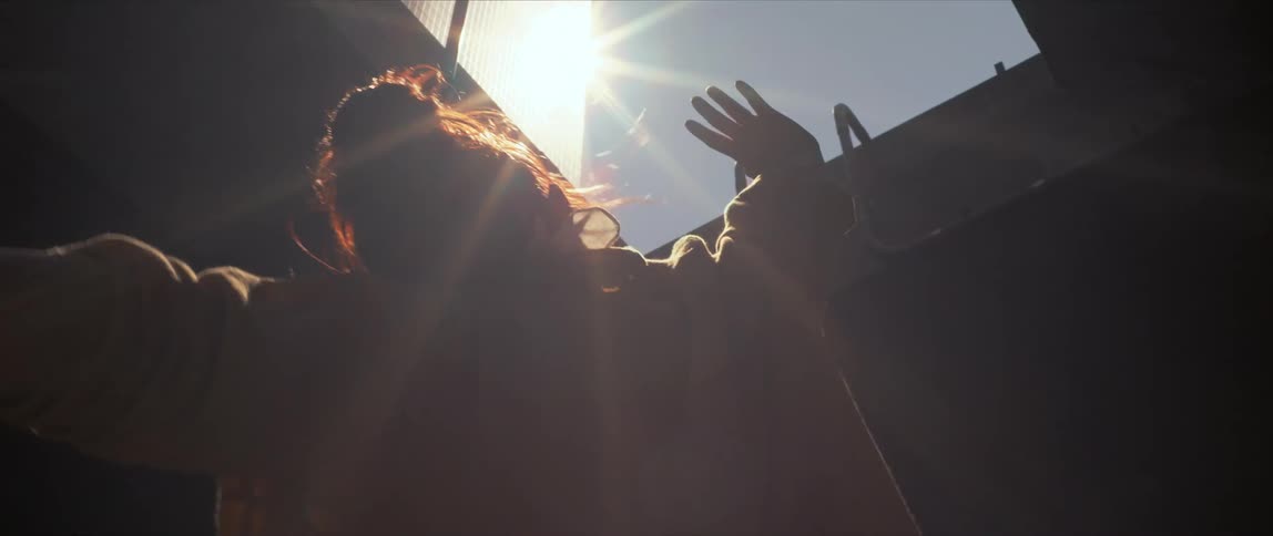 Sunlight shines through a gap in a building onto a dancing young woman in a medical mask