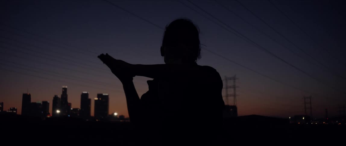 A woman dances outdoors in front of a city skyline with her hands clasped