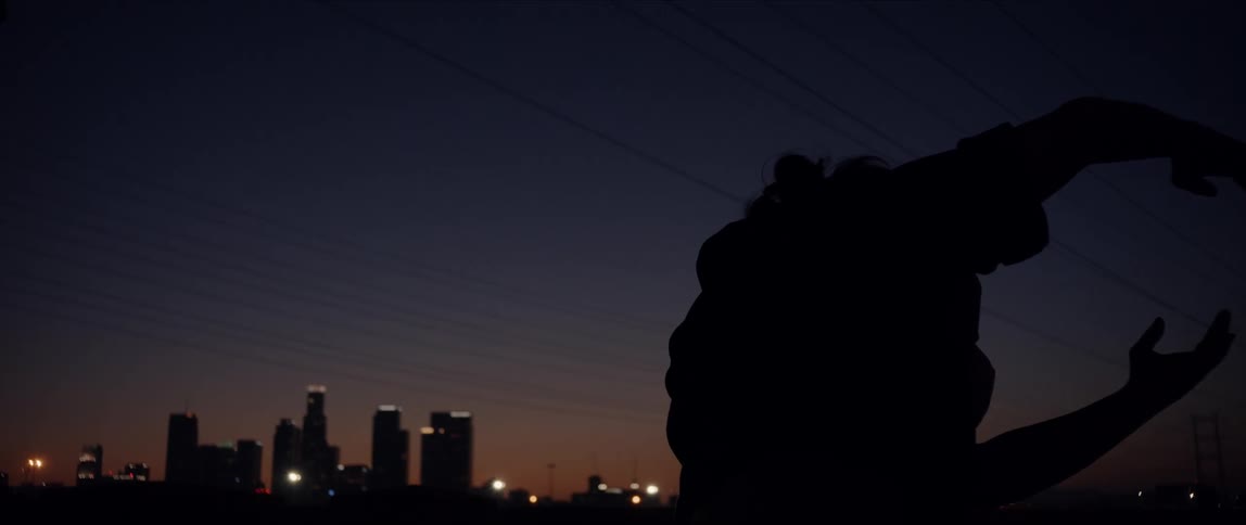 Silhouette of female dancer turns and moves under power lines at night with city skyline on horizon