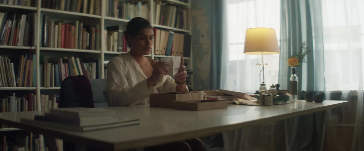 A young woman sitting at a table unfolds and reads a sheet of paper