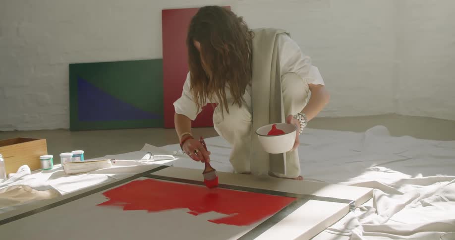 A woman uses orange paint to apply to a white canvas lying on the floor