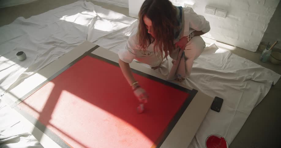 Woman dressed in white crouches down while painting red paint on a canvas