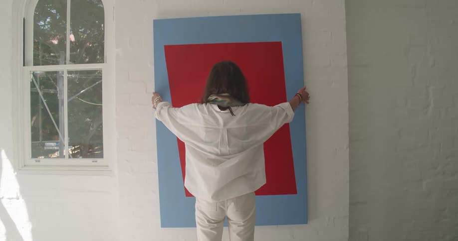 A woman holds up a large painting of a red square against a blue background in front of a white brick wall