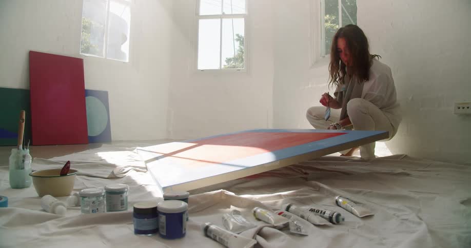 A woman squatting holding a piece of art work strokes blue paint on the edge in a room at daytime