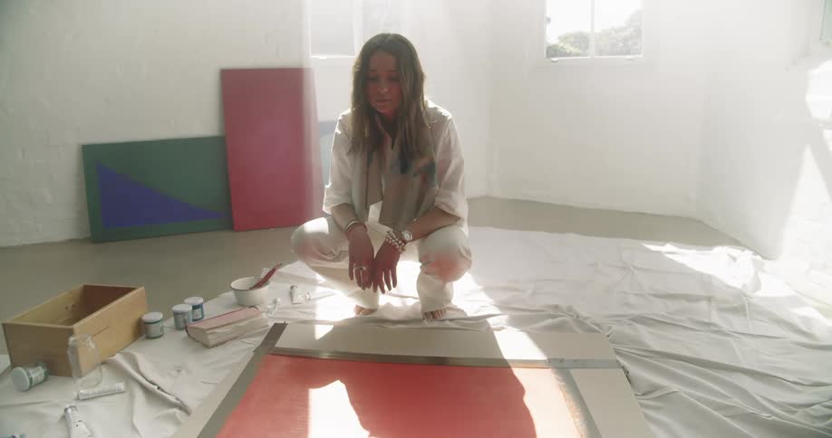 A woman crouching on the floor on a cloth tarp staring at a canvas painting