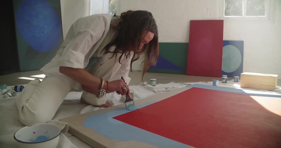 Woman kneeling on the floor paints a stroke of blue paint on a painting inside of an art studio