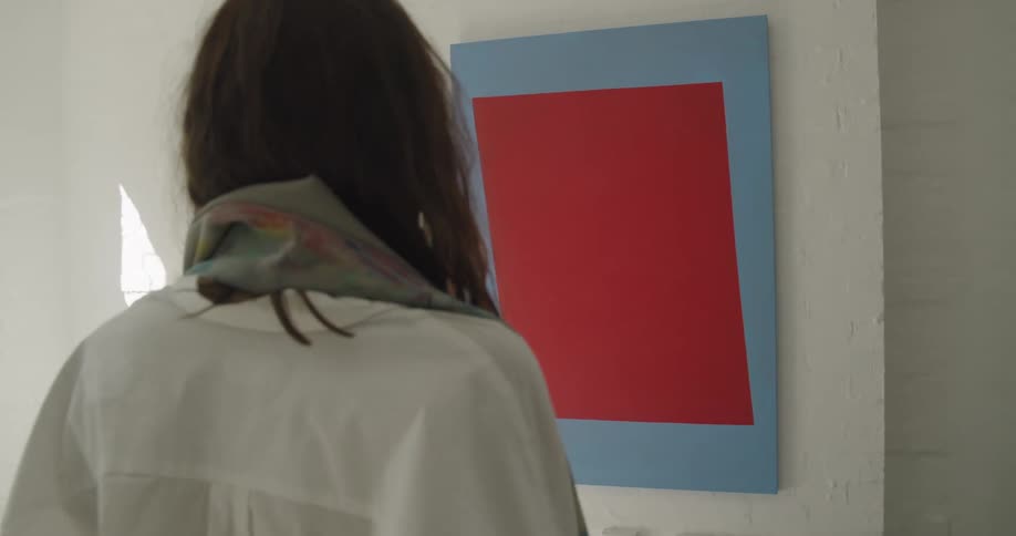 A young woman walks up to and looks at an abstract artwork hanging on the wall