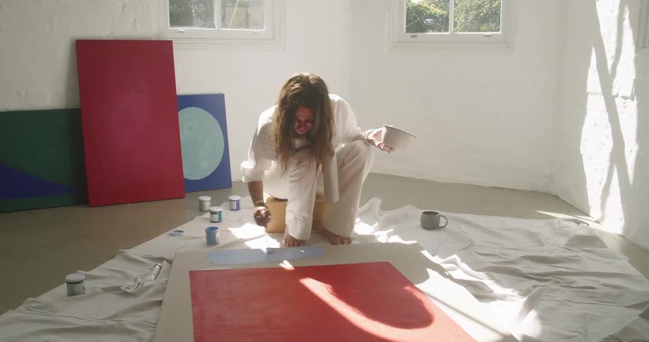 Woman using a large paintbrush to brush blue paint onto a piece of artwork on the floor in a room at daytime