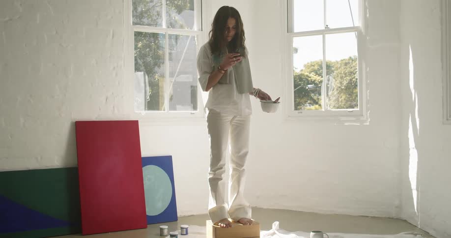 A woman stands on a box in the middle of a room while using a cell phone