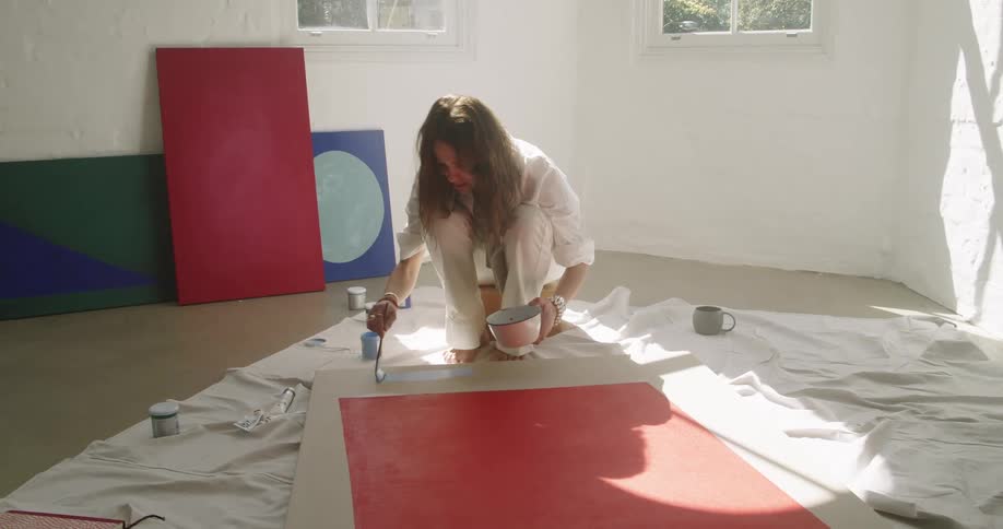 Woman kneeling down stroking light blue paint along the frame of an art piece of the floor in a room at daytime