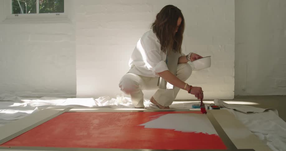 A woman kneeling in a room uses a paint brush to paint on a large canvas lying on a cloth on the floor of a room
