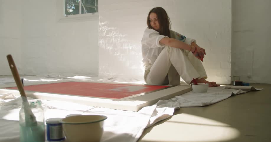 White woman sitting down next to her painting while looking at it