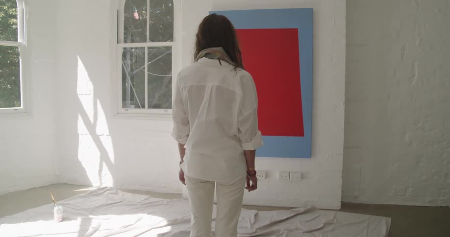 Woman stands in front of a portrait hung up on a wall in a room and stares at it at daytime