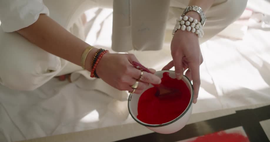 A person crouching on a cloth tarp stirring red paint in a bowl with a paintbrush