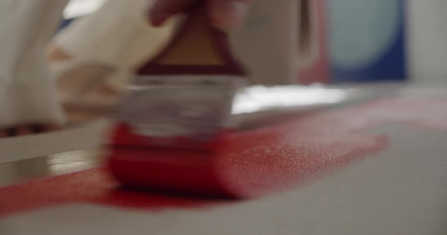 A person uses a broad paint brush to apply red paint to a pliable canvas