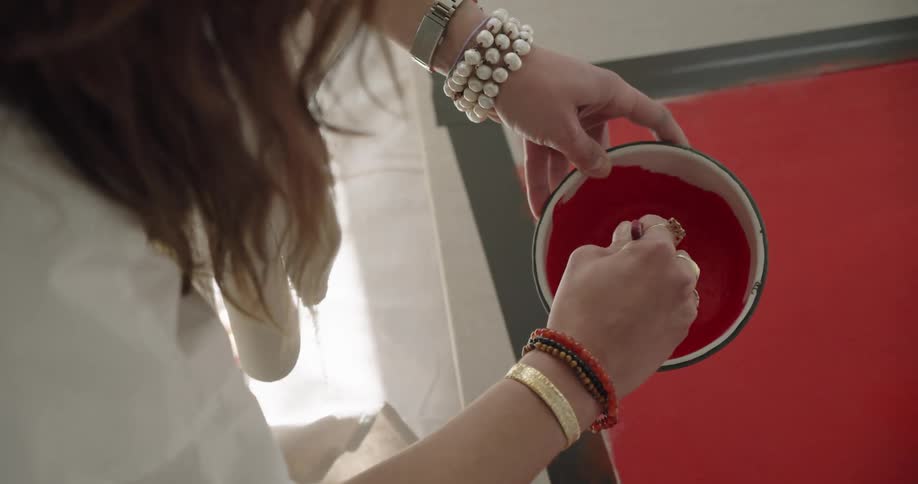 A woman stirs red paint in a bowl while standing over a large canvas on the floor
