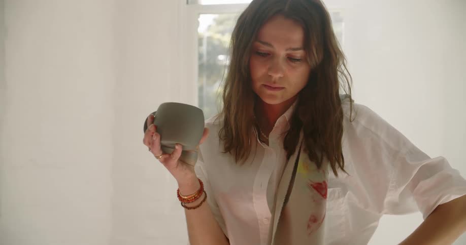 A woman stands indoors holding a coffee mug looking down and thinking