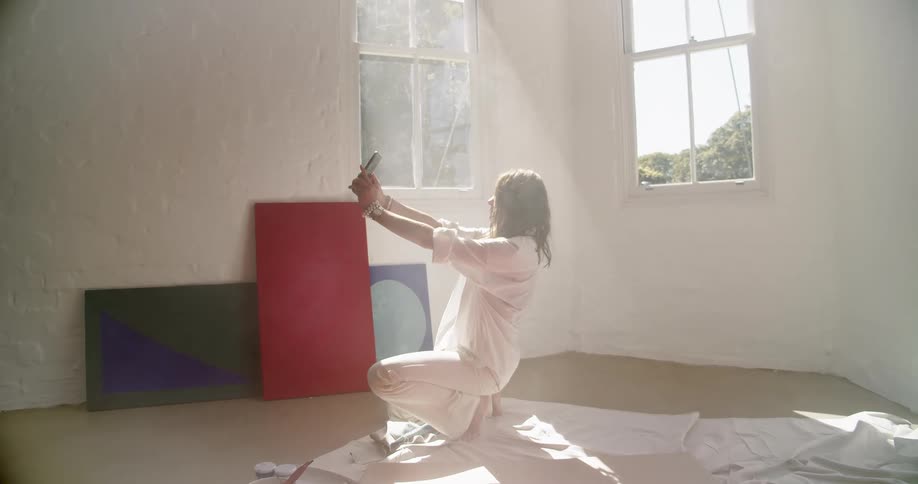 Woman crouches on the floor while holding a cellphone out in front of her as the sun streams through the window