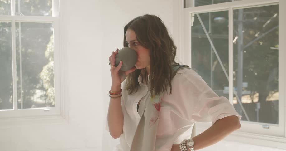 A woman standing lost in thought near a window in a house sips from a coffee mug while she thinks