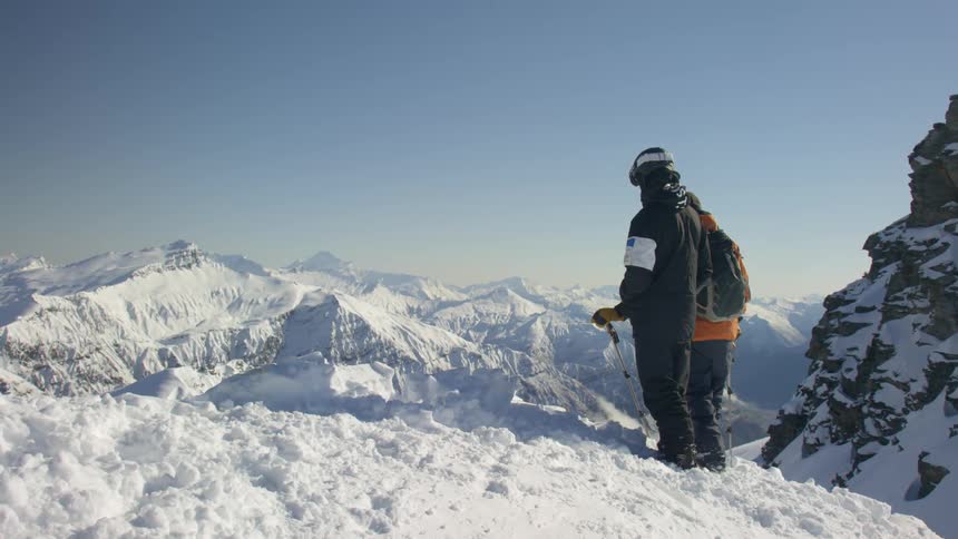 Two people standing at the top of a mountain and look down before skiing down it