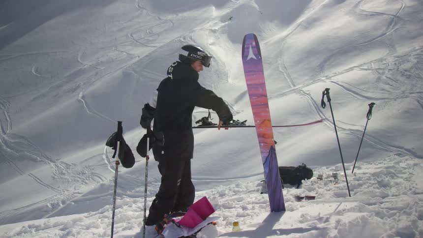 A man at the base of a mountain picks his skis up out of the snow and lies them down