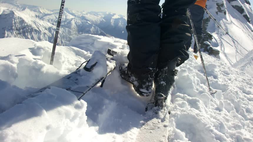 A pair of skiers stand next to one another on a mountain as one kicks snow off their boots