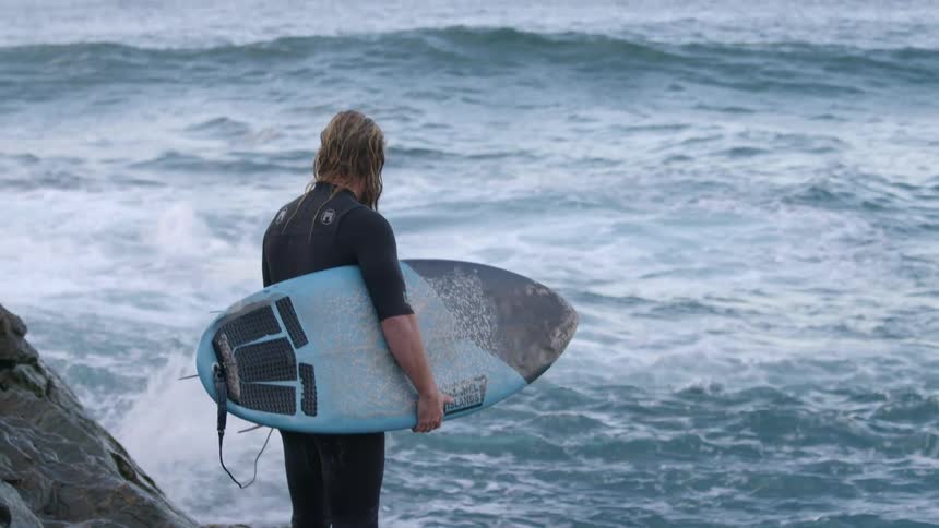 Man in a wet suit looks out at the ocean waves as he holds a body board