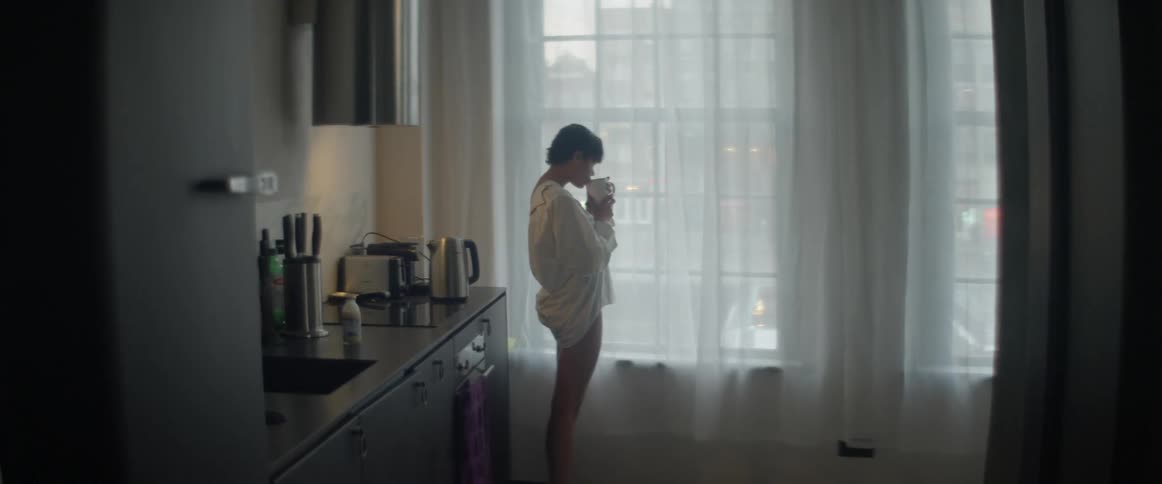 A woman standing in a kitchen drinking from a mug with windows in the background