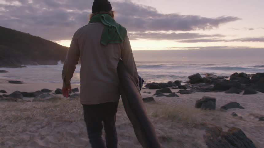 Man carrying a packed surfboard on the sands of a beach at sunset