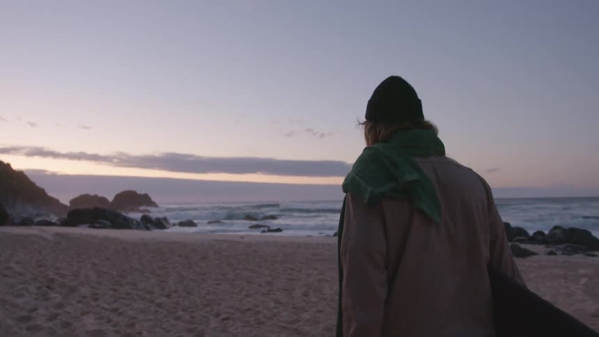 Man walks along beach carrying surfboard as sun tints sky on horizon