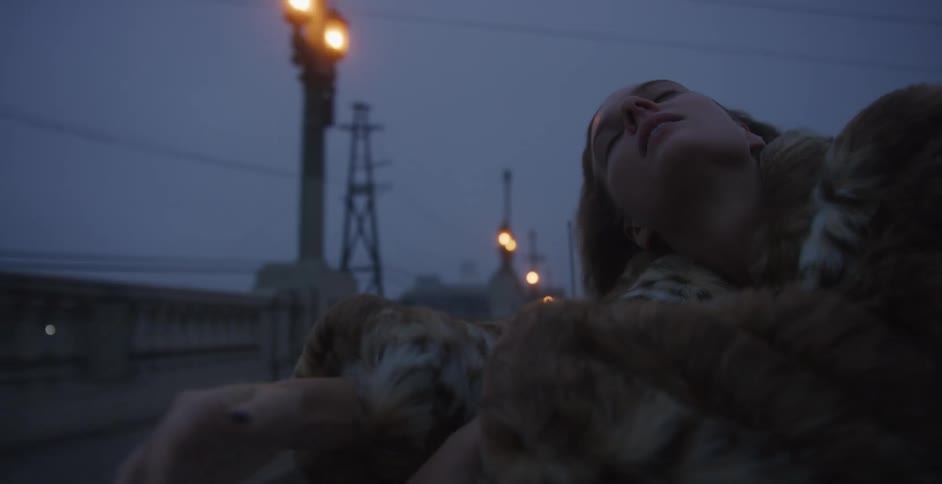 A woman wearing a fur coat sits outside on a moving vehicle travelling down a road