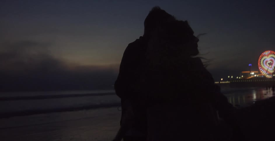A man and a woman hugging and walking on a beach with a ferris wheel the sky and clouds in the background