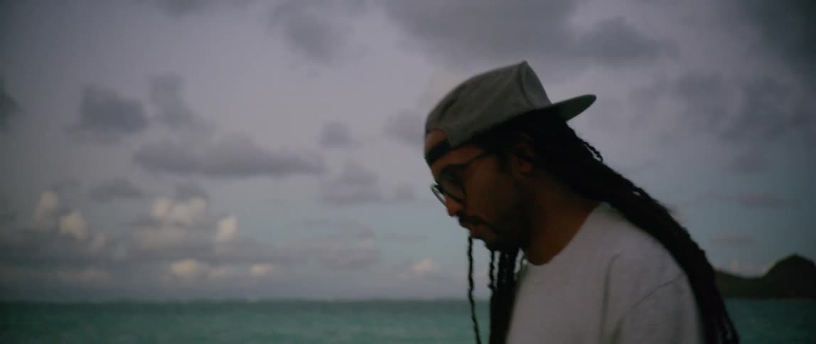 A man with long dreadlocks walks along the beach next to the water at dusk