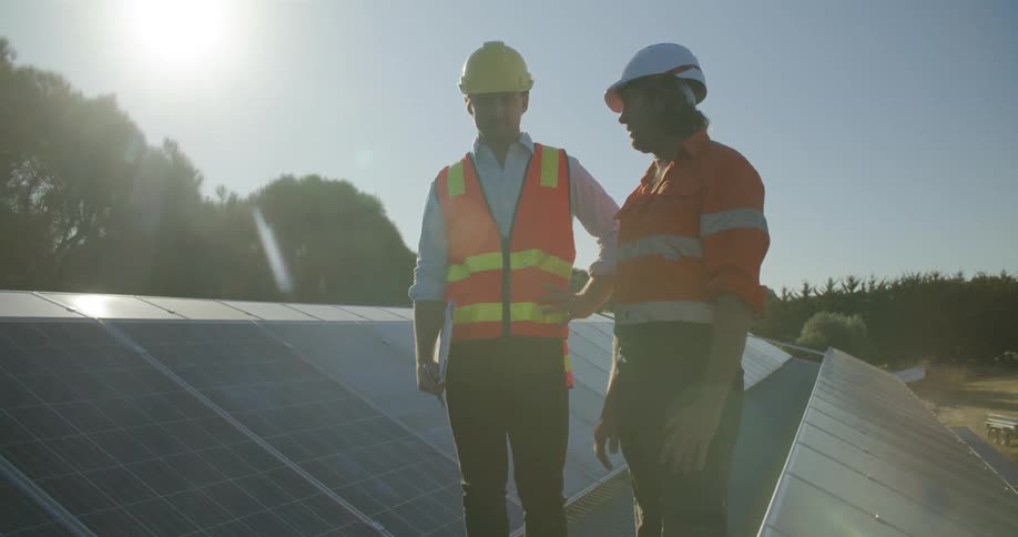 Two men in construction gear on a roof with solar panels at daytime talking to each other