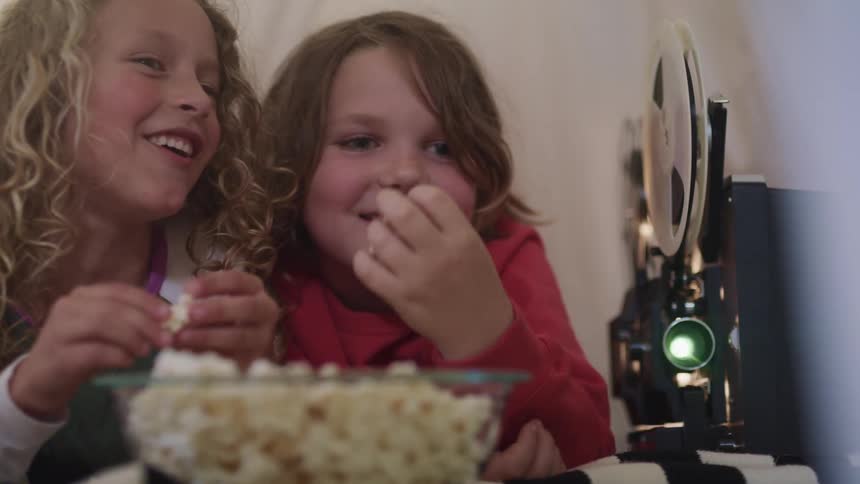 Two little girls laugh and talk as they eat popcorn and watch a movie at home
