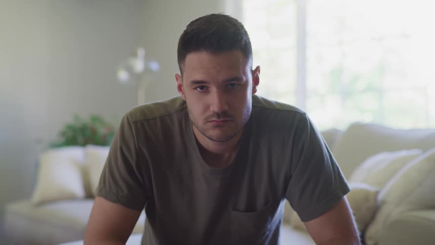 White man sitting down at his living room and staring at the camera