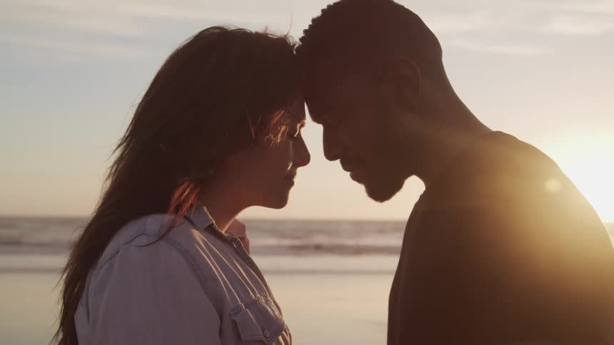 Man with beard and woman stand head to head with noses touching on the beach at sunset