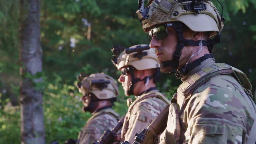 Military men wearing combat gear and holding rifles stand in line bordering tree and brush