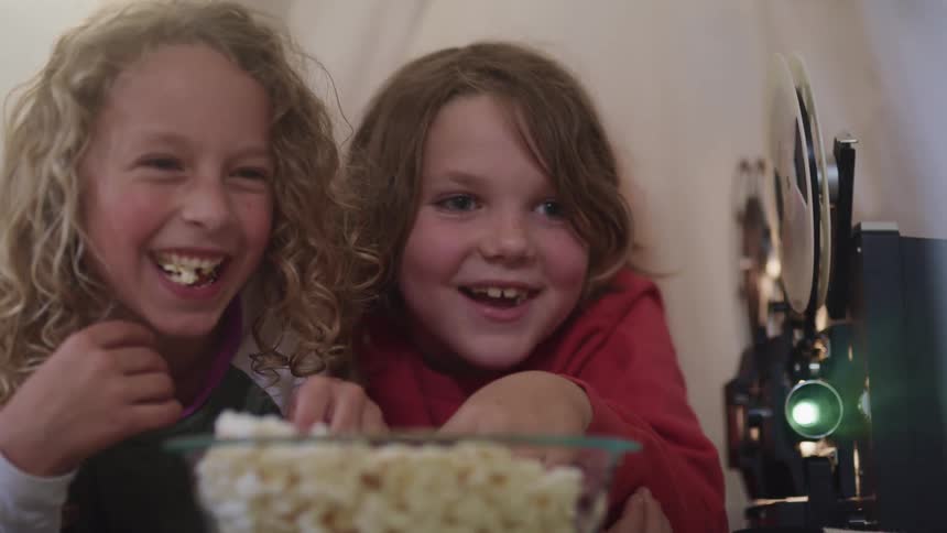 Brother and sister eating popcorn on a bed sheet tent watching movies on a vintage projector