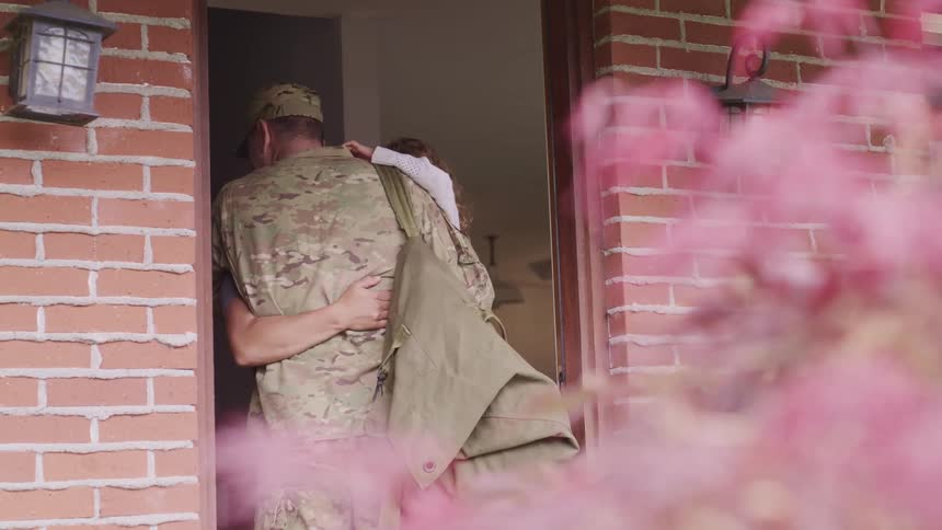 A woman hugging a man in military camouflage inside a house doorway