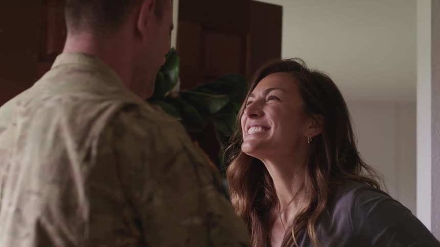 Woman smiles broadly at man in military fatigues hugging him in welcome as they stand near front door