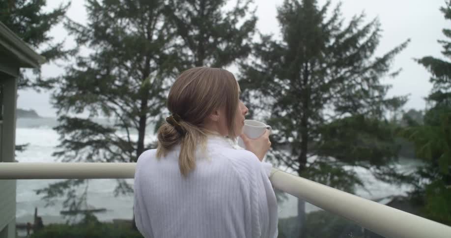 A woman holding a mug stands outside on a balcony overlooking beach