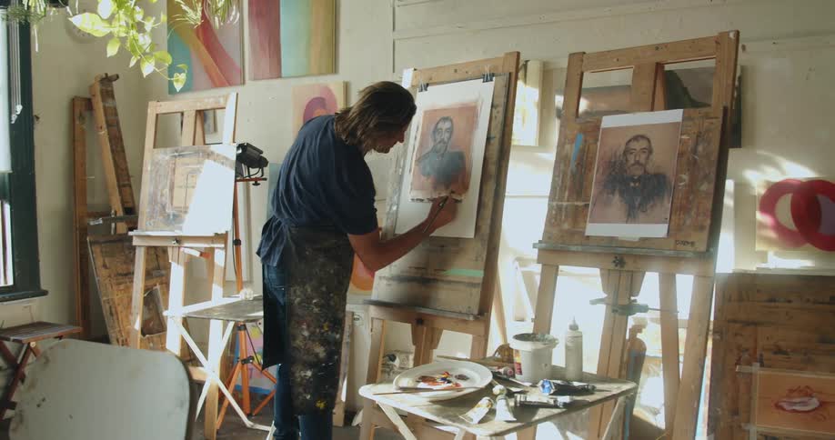 A man paints on a canvas surrounded by other paintings and wood frames on easels