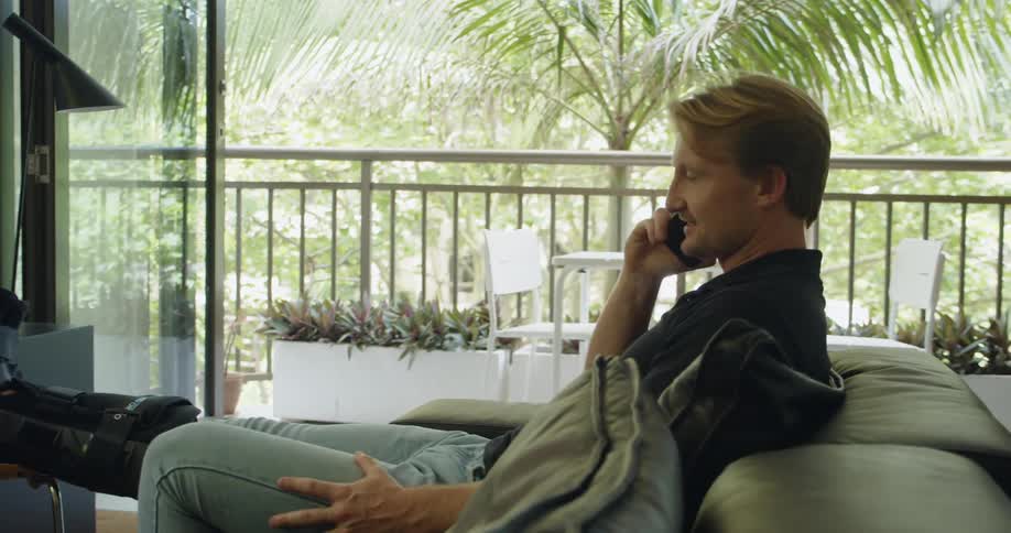 A man with a cast sitting down on a couch talking on a smart phone