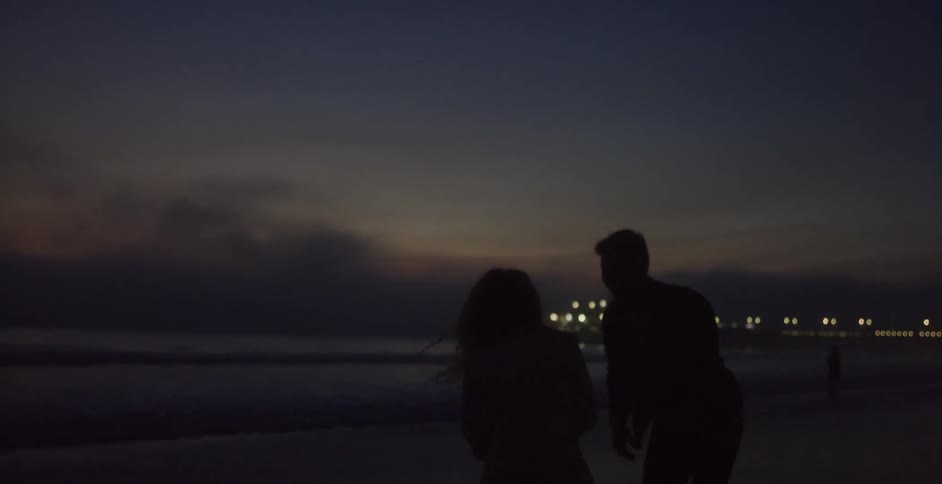 A man and woman playing and spinning around on the beach at night with the city skyline lit up in the distance
